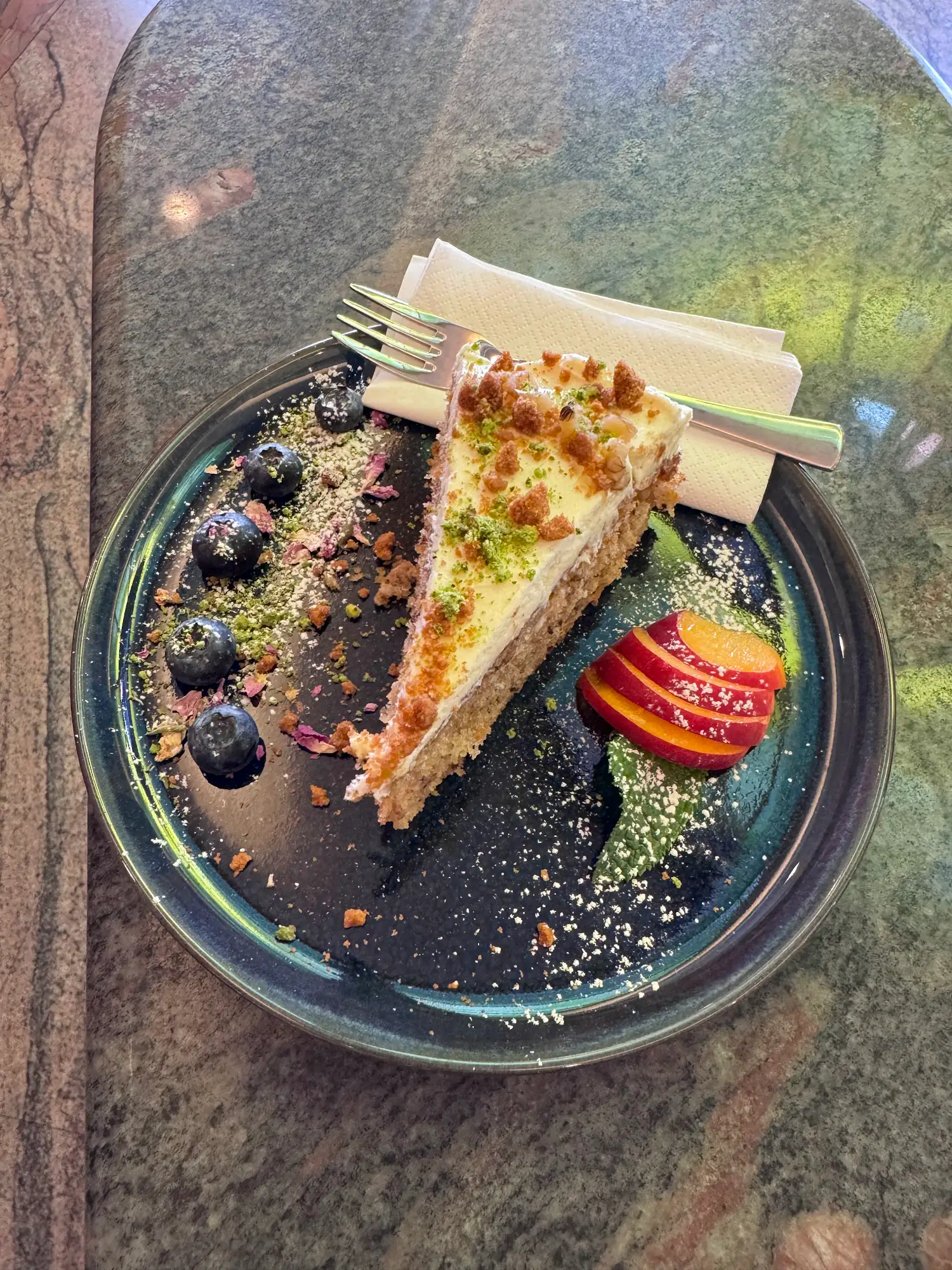 Tortenschnitte mit Pistazien, Heidelbeeren und Zwetschkenspalten auf grünem Marmortisch